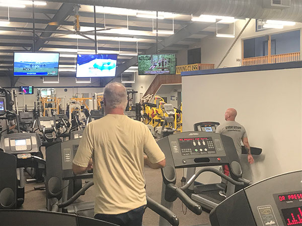 a modern gym with 2 men walking on different treadmills both looking at tv display screens